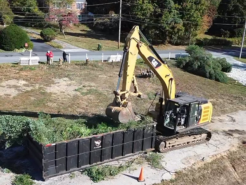 Excavator loading cleared brush into a roll-off container on a residential lot