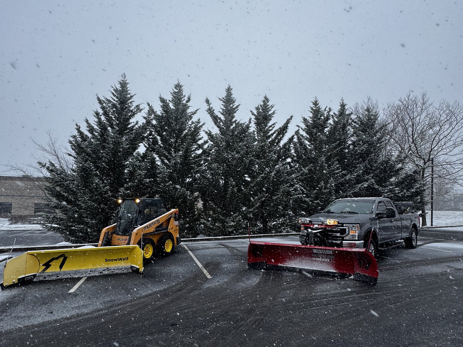 Plow truck and skid-steer snow pusher clearing a commercial parking lot during a snowfall